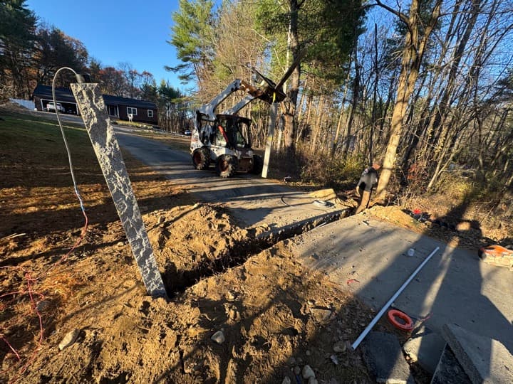 Underground utility trench cut along a private driveway with a Bobcat staged in the background, conduits prepared for service entrance.