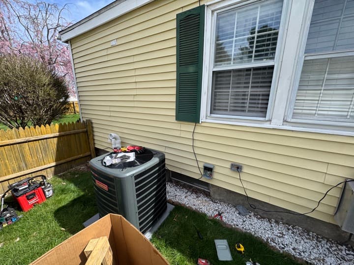 Residential central A/C condenser installed on a pad with disconnect mounted nearby, viewed from the yard.