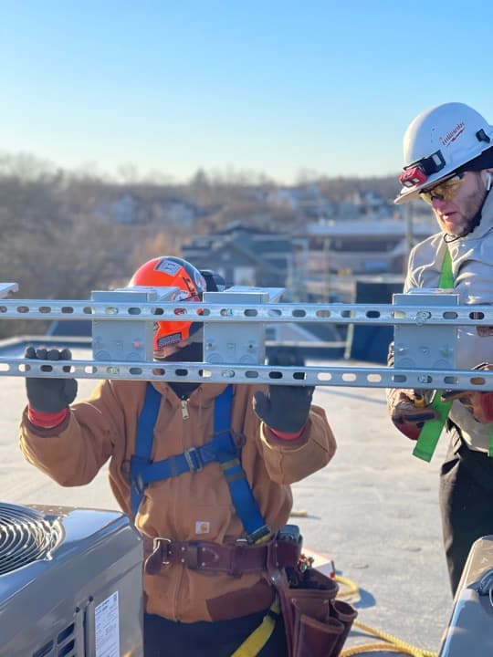 Brian holding strut steady while a coworker drills mounting hardware on a commercial rooftop.
